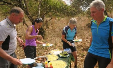 Group of four cyclists serving themselves food from a table in a natural setting.