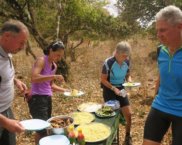 Group of four cyclists serving themselves food from a table in a natural setting.
