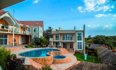 A curved blue swimming pool sits between orange and beige resort buildings, with thatched roofs and a bright sky above.