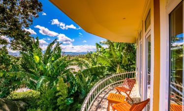 A curved balcony with orange wooden chairs overlooks dense banana trees and a bright blue sky with scattered clouds.