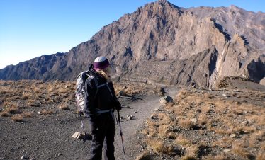 Hiker standing on a trail with a mountain in the background, wearing a backpack and using trekking poles.