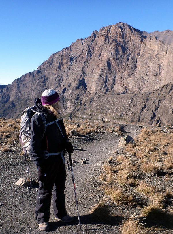 Hiker standing on a trail with a mountain in the background, wearing a backpack and using trekking poles.