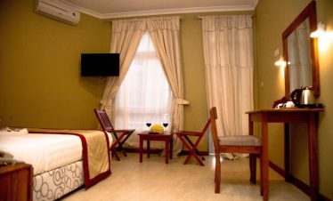 A neatly arranged hotel room has a bed, two wooden chairs, a small table, a wall-mounted TV and a kettle on a side desk.
