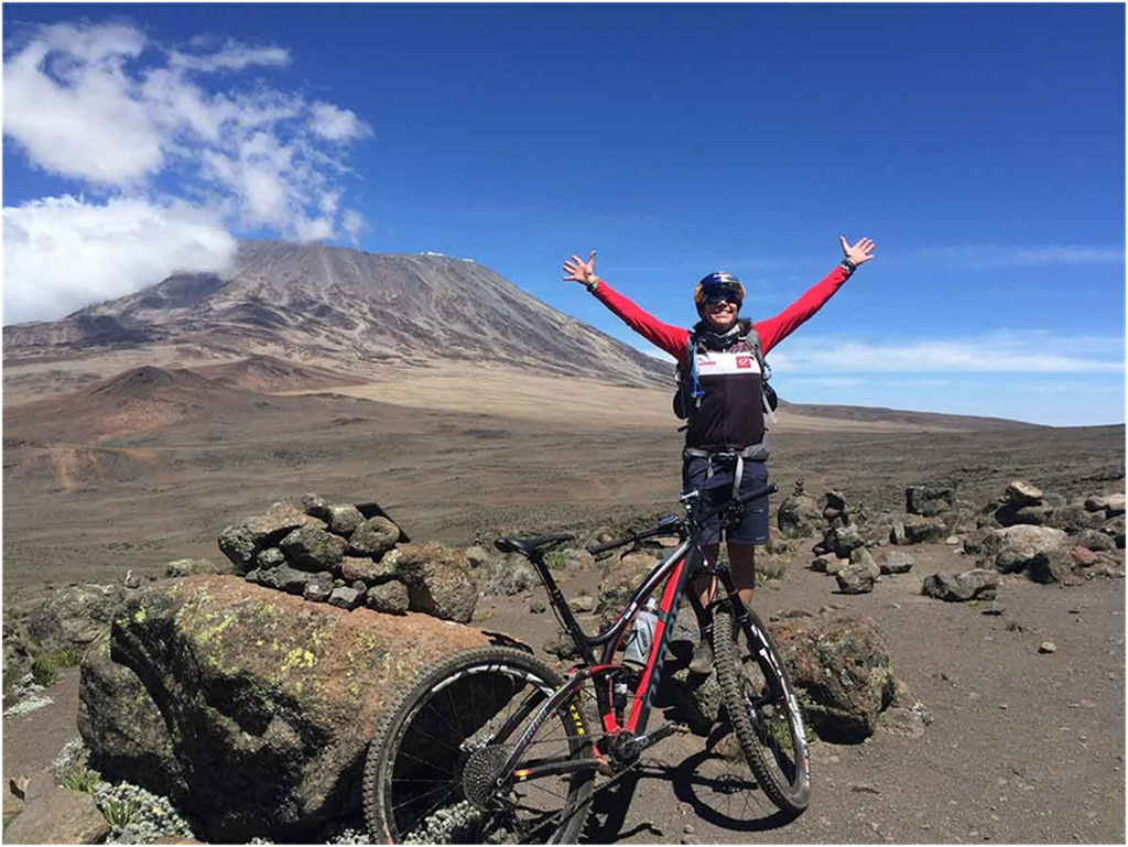 Individual with arms raised standing next to a mountain bike with Mount Kilimanjaro in the background.