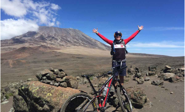 Individual with arms raised standing next to a mountain bike with Mount Kilimanjaro in the background.