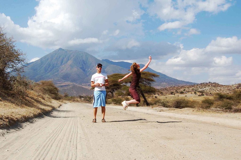 A man standing with a woman jumping joyfully on a dirt road with a mountain backdrop.