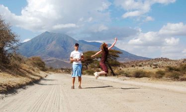 A man standing with a woman jumping joyfully on a dirt road with a mountain backdrop.