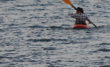 Individual paddling a red kayak with an orange paddle on a body of water.