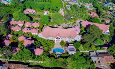 An aerial view shows a tropical lodge compound with red-roofed buildings, a central swimming pool, and dense green gardens.