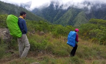 Two hikers wearing colorful backpacks walking through a grassy area with mountains in the background.