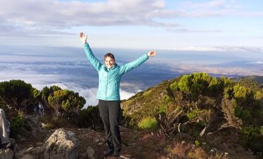 Individual wearing a blue jacket standing with arms raised on a mountain peak overlooking a scenic landscape.