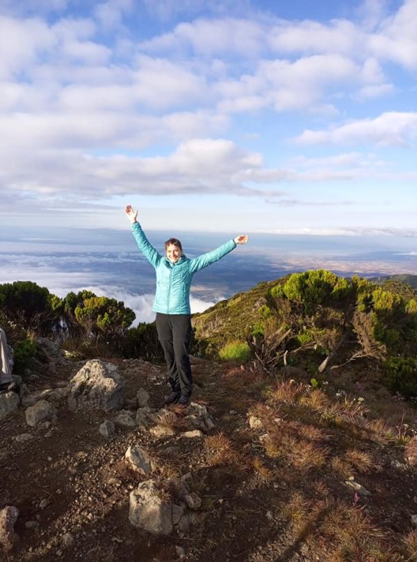 Individual wearing a blue jacket standing with arms raised on a mountain peak overlooking a scenic landscape.