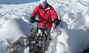 Individual in a red jacket and colorful hat maneuvering a bicycle through snow-covered terrain.