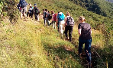 A group of hikers walking along a mountain trail surrounded by greenery and hills.