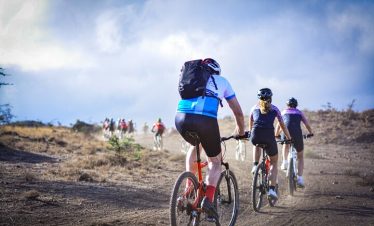 Group of mountain bikers riding on a dirt trail through a hilly landscape under a cloudy sky.