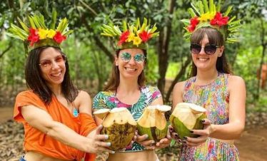 Smiling women wearing floral crowns hold freshly opened coconuts during a tropical outing in a leafy forest clearing with bright green foliage.