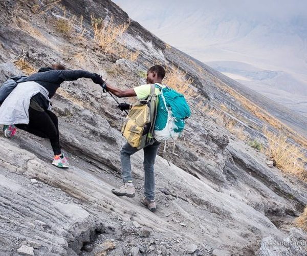 Two climbers navigating a steep rocky slope, one assisting the other with a handhold.