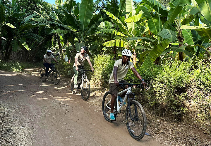 Three individuals riding bicycles along a dirt path surrounded by banana plants in Mto Wa Mbu.