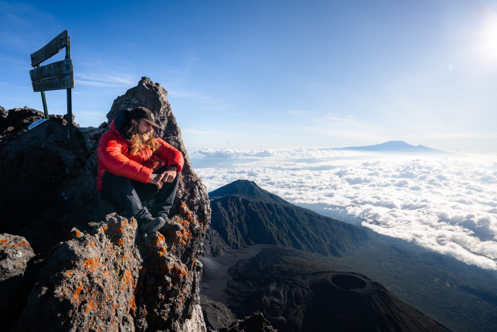 Individual in a red jacket sitting on a rocky outcrop at Mount Meru, overlooking clouds and distant mountains.