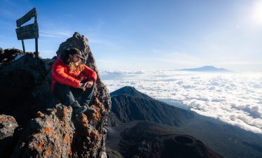 Individual in a red jacket sitting on a rocky outcrop at Mount Meru, overlooking clouds and distant mountains.