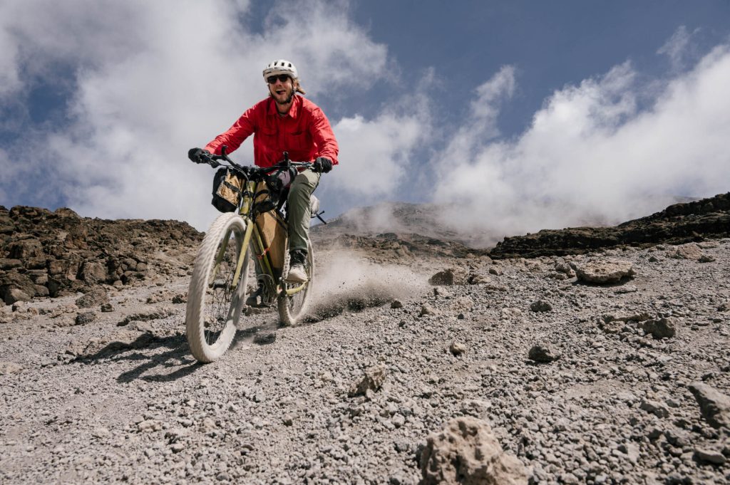 A cyclist in a red jacket riding a mountain bike on rocky terrain with clouds in the background.