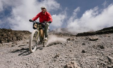 A cyclist in a red jacket riding a mountain bike on rocky terrain with clouds in the background.
