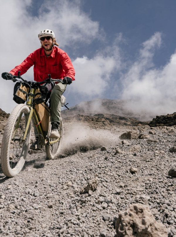 A cyclist in a red jacket riding a mountain bike on rocky terrain with clouds in the background.