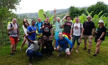 A group of hikers poses outdoors near Materuni village with lush green banana plants and rolling hills under overcast skies around Kilimanjaro.