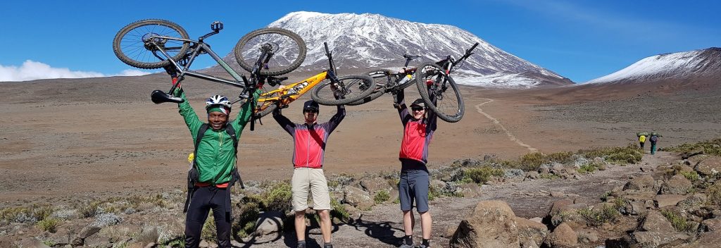 Three mountain bikers holding their bikes above their heads in front of Mount Kilimanjaro with snow-capped peak.