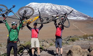 Three mountain bikers holding their bikes above their heads in front of Mount Kilimanjaro with snow-capped peak.