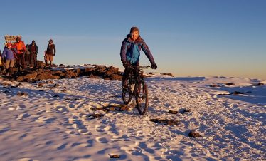 A mountain biker standing on snow-covered terrain at the summit of Kilimanjaro with a group of hikers in the background.