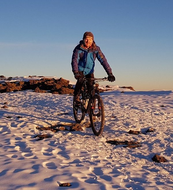 A mountain biker standing on snow-covered terrain at the summit of Kilimanjaro with a group of hikers in the background.