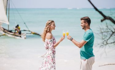 On a tropical shoreline, a woman in a floral dress and a man in a turquoise T-shirt toast with champagne as a dhow sails past.