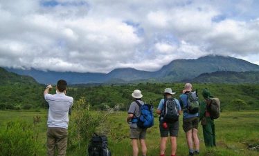 Hikers with backpacks stand on a trail under a cloudy sky, taking in lush green plains and distant mountainous silhouettes.