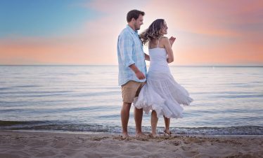 A couple stands barefoot on the beach at sunset, with gentle waves, a pastel sky, and a flowing white dress in the breeze.