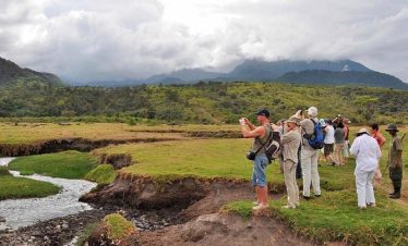 Hikers with backpacks pause along a grassy trail, looking over a green valley and distant mountainous ridges under a cloudy sky.