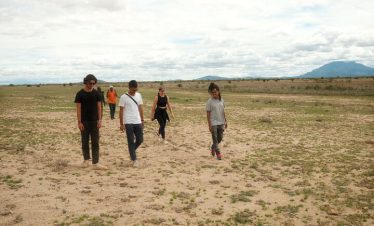 A group of hikers walks across a dry open savannah under cloudy skies, with distant hills and mountains on the horizon behind them.