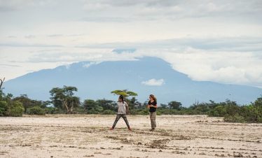 Two hikers walk across dusty open ground with vast greenery and a mountainous horizon under layered clouds in the distance.