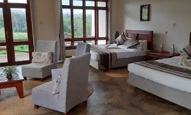 A bright lodge room with two twin beds, cushioned chairs, wooden tables and large windows looking out onto green hills and trees outside.
