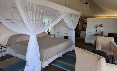 A spacious safari lodge tent room with a four-poster bed draped in mosquito netting, beige seating, and a desk beside the window.