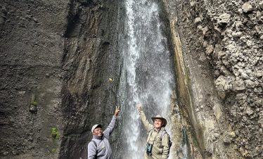 Two hikers pose with arms raised beneath a cascading waterfall inside a rocky canyon, surrounded by wet stones, mist, and greenery.