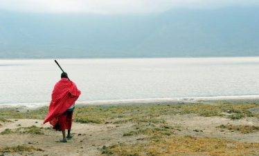 Maasai individual wearing a red shuka standing by the water's edge with a stick in hand.