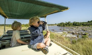 Sitting on a boat under a green canopy, a child peers through binoculars while another child looks on as a river flows by.