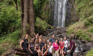 A group stands near the Materuni waterfalls in Tanzania, surrounded by lush green rainforest trees and rocky terrain.