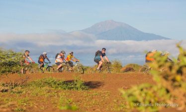 A group of cyclists riding on a dirt path with a mountain in the background under a clear sky.
