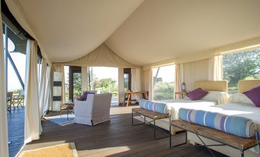 Inside Mara Mara Tented Lodge, a spacious safari tent suite offers twin beds, lounge chairs and wide views through large windows across the savanna.