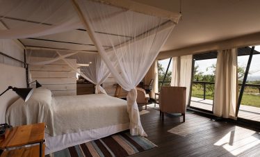Inside Mara Mara Tented Lodge, a safari tent bedroom with a canopy bed, mosquito netting, wooden floors and a deck overlooking the savanna outside.