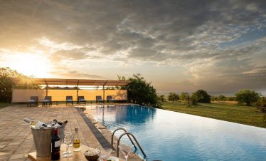 At Mara Mara Tented Lodge, an infinity pool overlooks the Serengeti plains at sunset, with lounge chairs, drinks, and a covered deck nearby.