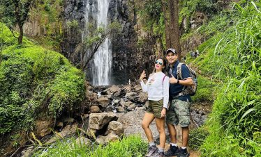 A bright waterfall at Materuni plunges over rocky cliffs, framed by lush green vegetation and two hikers posing on a trail.