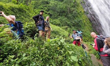 Travelers on a hike through lush green forest near the Materuni waterfall, with hikers taking photos as water streams down.